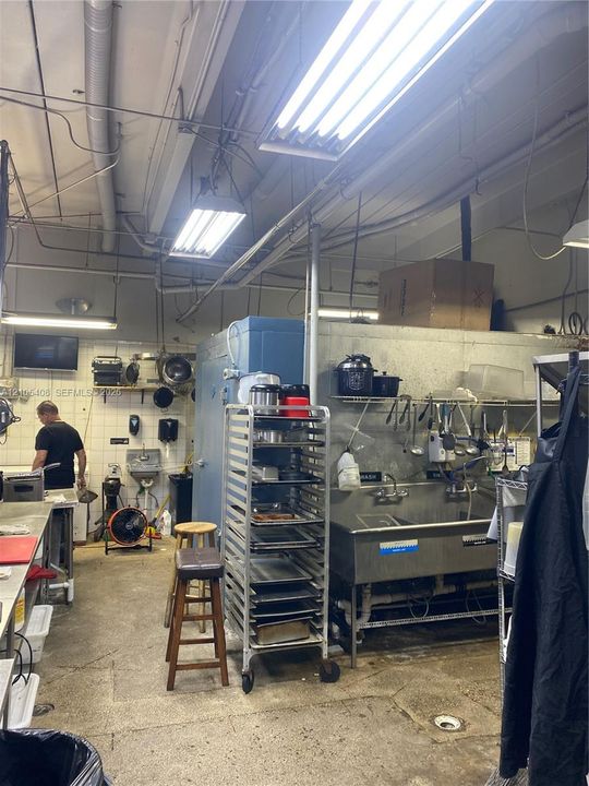 Commercial-style kitchen featuring multiple fluorescent light fixtures, exposed ductwork, a stainless steel sink with mounted shelving, and a large reach-in refrigerator unit