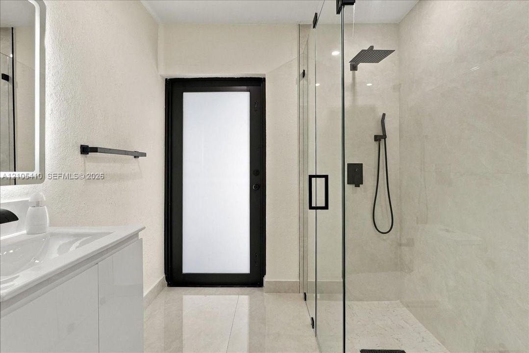 Bathroom featuring a walk-in shower with frameless glass enclosure, matte black rainfall showerhead, and handheld wand, accompanied by a white vanity with integrated sink and a frosted glass exterior door