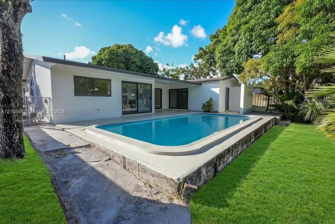 In-ground swimming pool with a concrete deck, sliding glass doors, and a grass lawn with mature trees