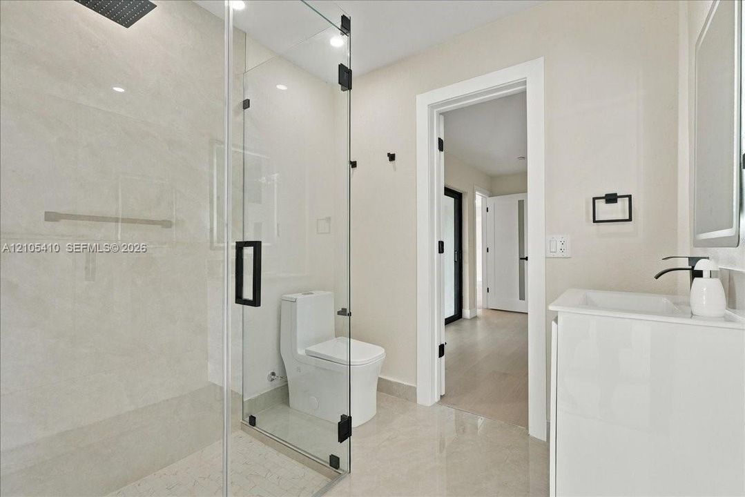 Frameless glass-enclosed shower featuring large-format tile surround, rainfall showerhead, sleek white vanity with integrated sink, and matte black fixtures