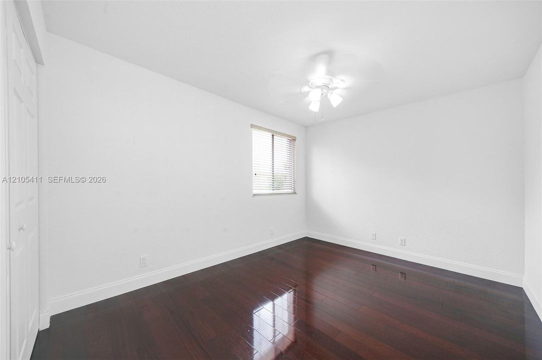 Dark wood-finish flooring, white walls, built-in closet with bi-fold doors, single window with horizontal blinds, and a ceiling fan with light fixture
