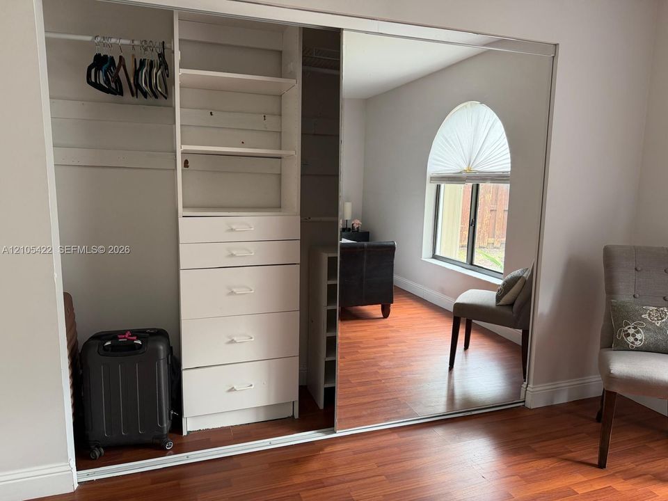 Built-in wardrobe featuring mirrored sliding doors, integrated shelving, and a multi-drawer storage unit, complemented by wood-finish flooring and an arched window
