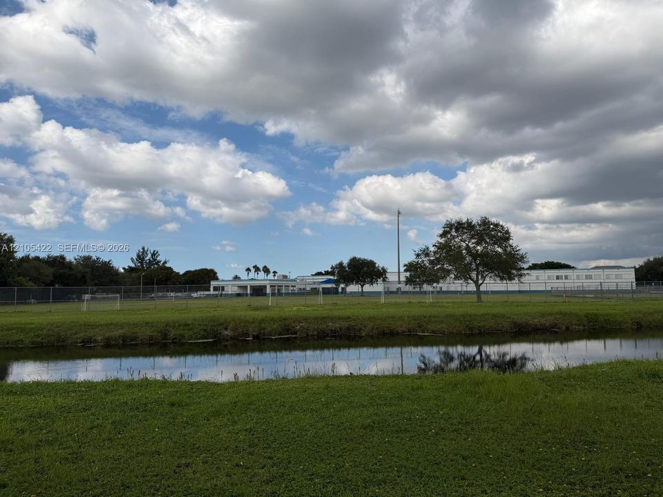 Open green space featuring a natural water element, perimeter fencing, and mature trees