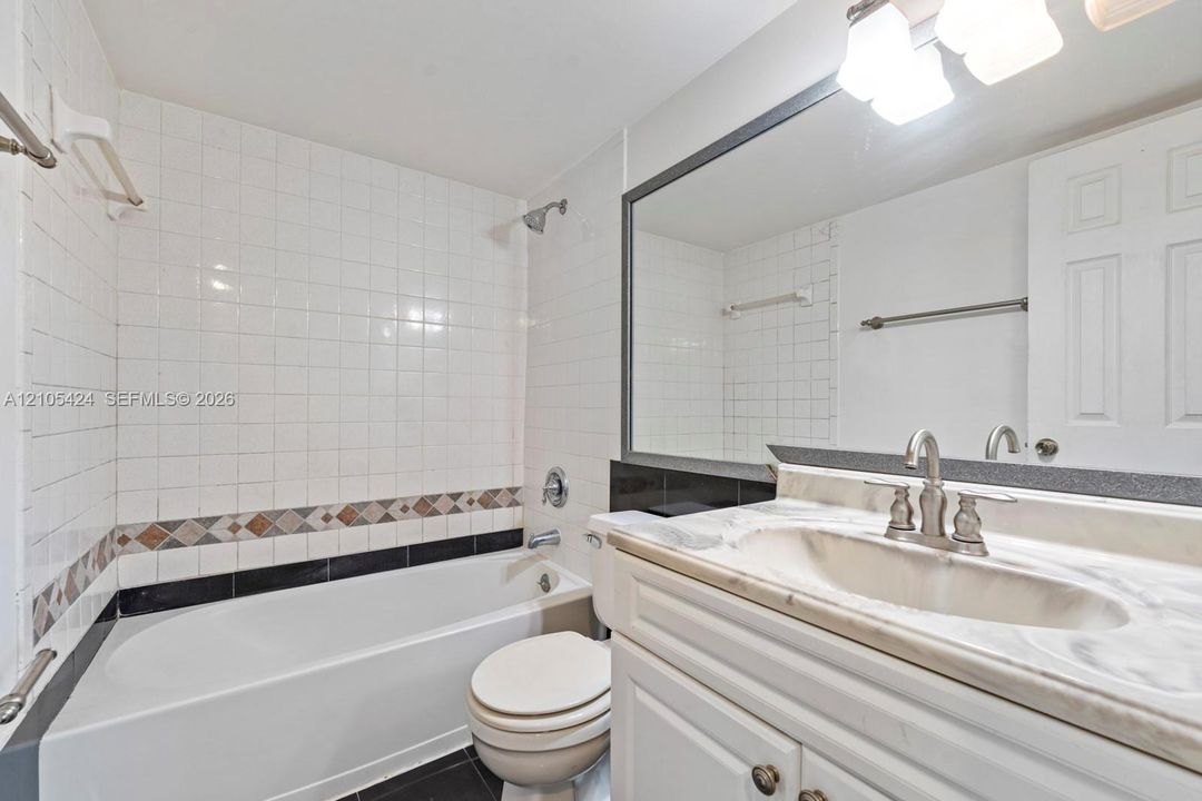 White tile shower surround with decorative accent band, tub with shower fixture, and a vanity with a white countertop and integrated sink