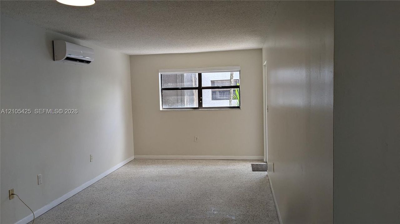 Wall-mounted air conditioning unit, light-colored walls, window with dark framing, textured ceiling, and light-colored carpeting