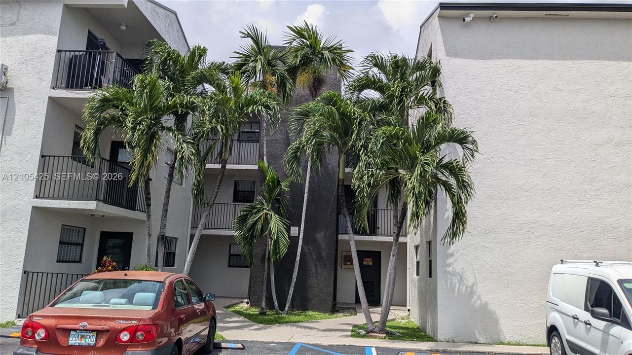 Multi-story building featuring balconies with black metal railings, textured light-colored stucco exterior, and mature palm trees