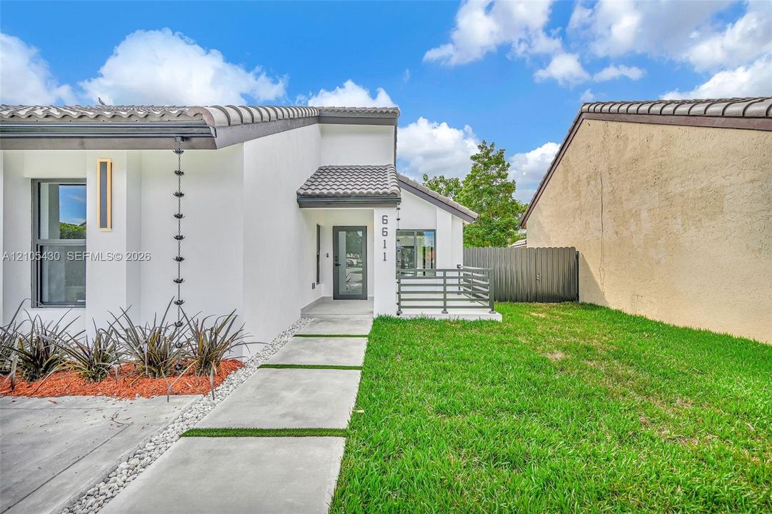 Exterior featuring smooth white stucco finish, concrete paver walkway with turf inlay, tile roof, vertical metal rain chain, and front patio with horizontal railing