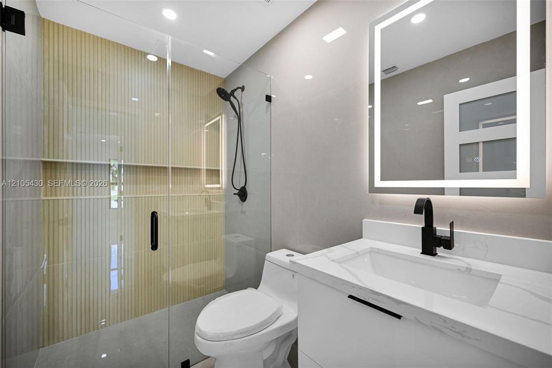 Bathroom featuring a floating vanity with a stone-top integrated sink, matte black hardware, an illuminated mirror, and a glass-enclosed shower with vertical-tile detailing
