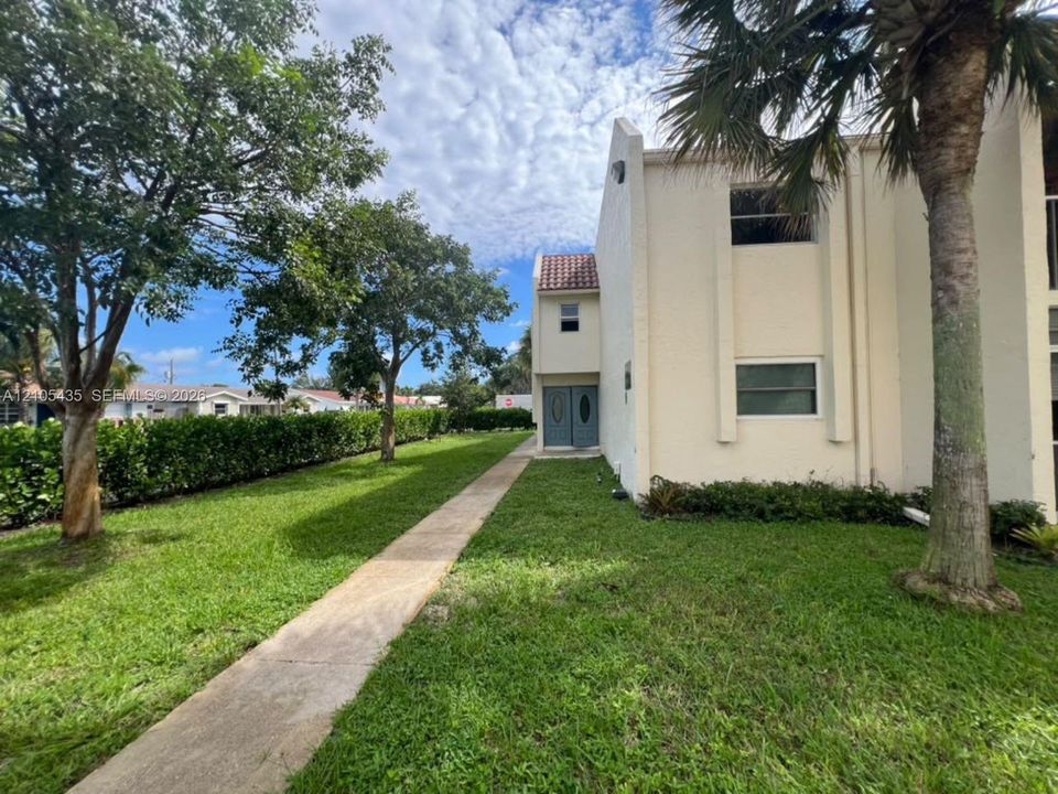 Exterior facade featuring light-toned masonry, a concrete paver walkway, double-door entryway, and mature palm trees situated on a grassy lawn