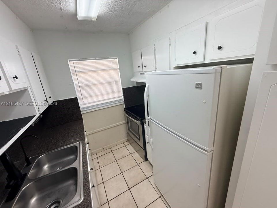 Dual basin stainless steel sink, black countertop, white cabinetry with black hardware, tile flooring, and integrated white appliances