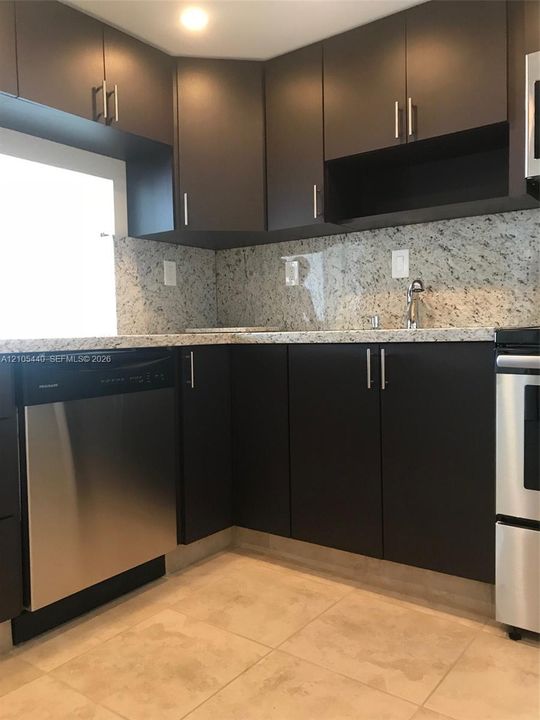 Dark wood-finish cabinetry with stainless steel hardware, light-toned stone-finish countertops and backsplash, stainless steel appliances, and large square floor tiles