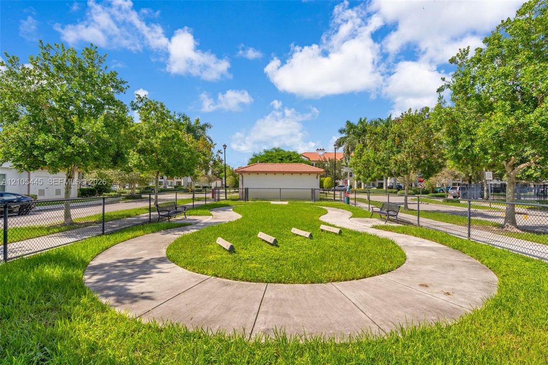 Fenced outdoor recreation area featuring a concrete walking path, manicured lawn, perimeter benches, and mature trees