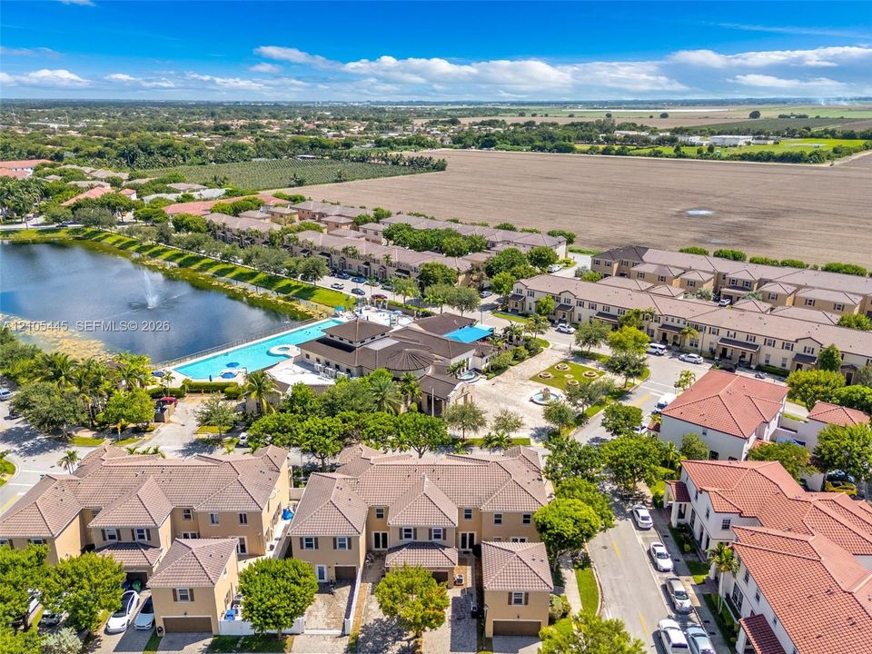 Aerial perspective of a multi-unit residential community featuring clay tile roofing, a large central swimming pool with an integrated fountain, paved pedestrian walkways, and mature landscaping