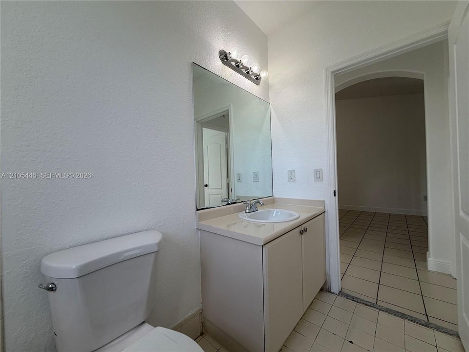 White toilet with chrome flush lever, vanity with integrated sink and chrome faucet, vanity mirror with overhead light fixture, and tile flooring