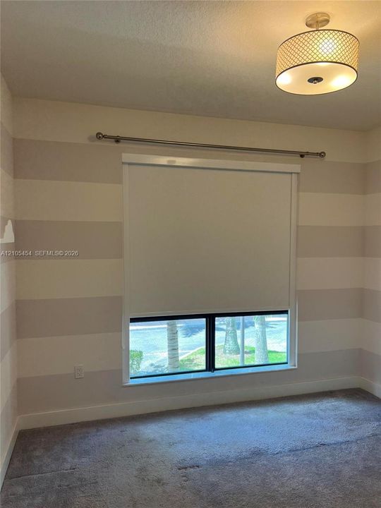 Striped wall treatment, carpeted flooring, window with roller shade, ceiling-mounted light fixture, and window rod