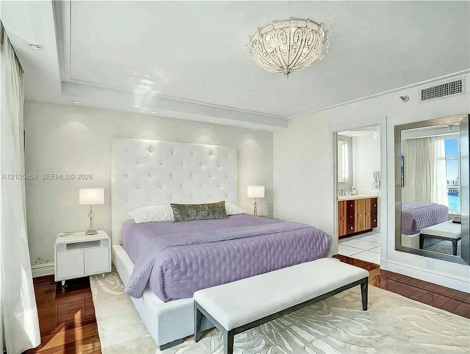 Tray ceiling with recessed lighting, ornate ceiling fixture, wood-finish flooring, tufted headboard, and an en-suite bathroom with a wood-finish vanity