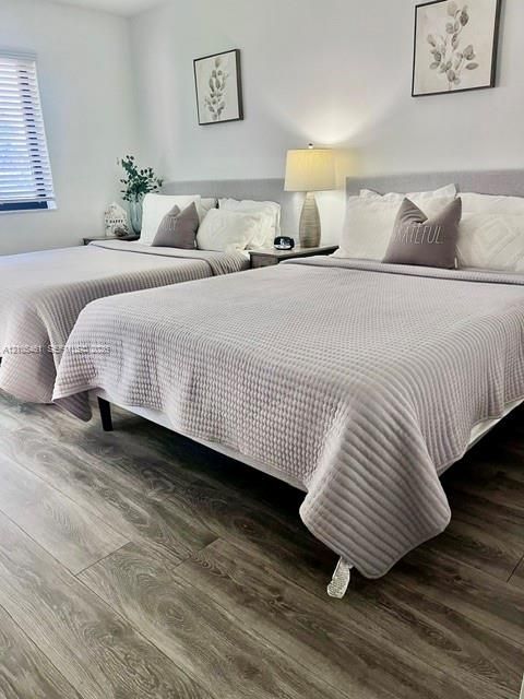 Wood-finish flooring, two beds with upholstered headboards, a window with horizontal blinds, and a built-in wall lamp