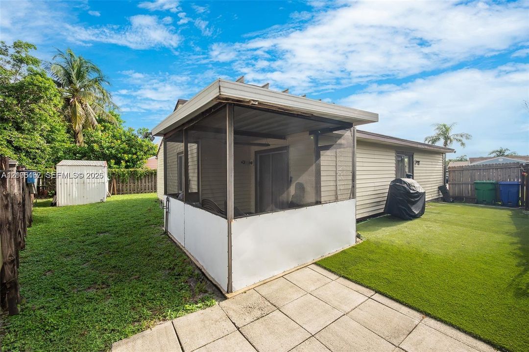 Screened-in patio with light-colored siding, concrete paver flooring, synthetic turf lawn, and detached storage shed