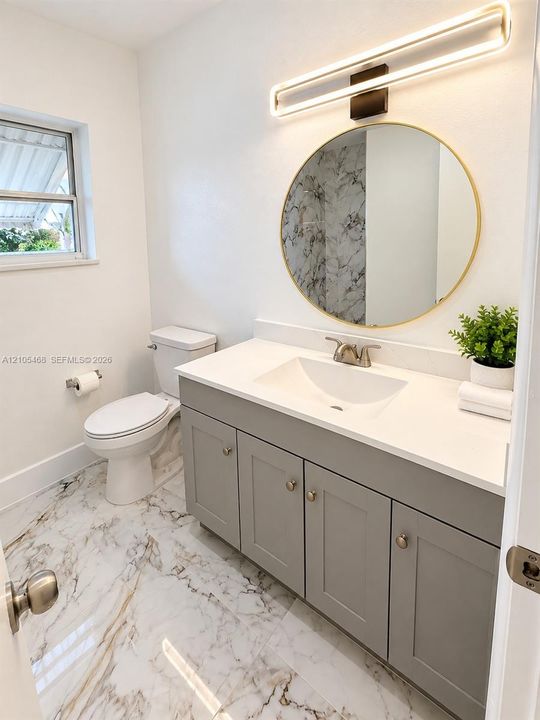 Vanity with a white countertop and integrated sink, gray cabinetry with brass-finish hardware, a circular mirror, and a wall-mounted linear light fixture