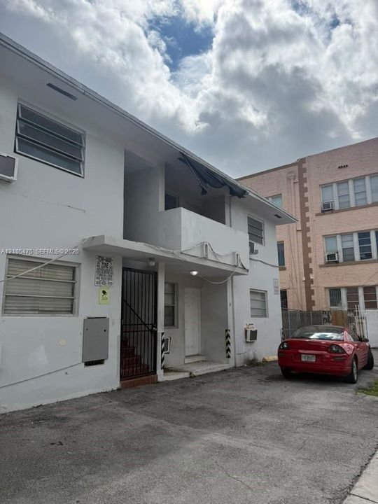 Two-story multi-unit structure featuring white stucco siding, louvered windows, a ground-level gated entryway, and a second-story balcony