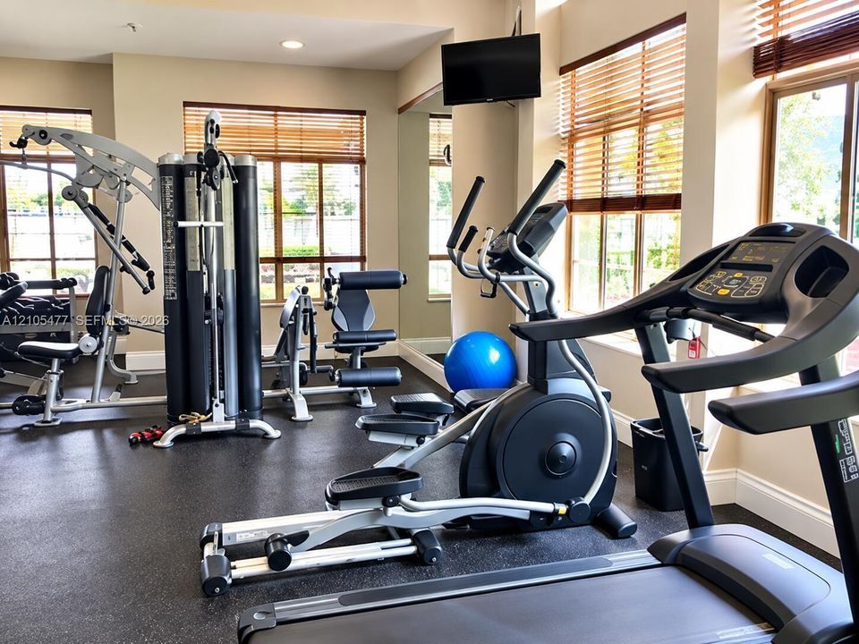 Dedicated fitness area featuring rubberized flooring, large windows with wood-finish blinds, and a wall-mounted display
