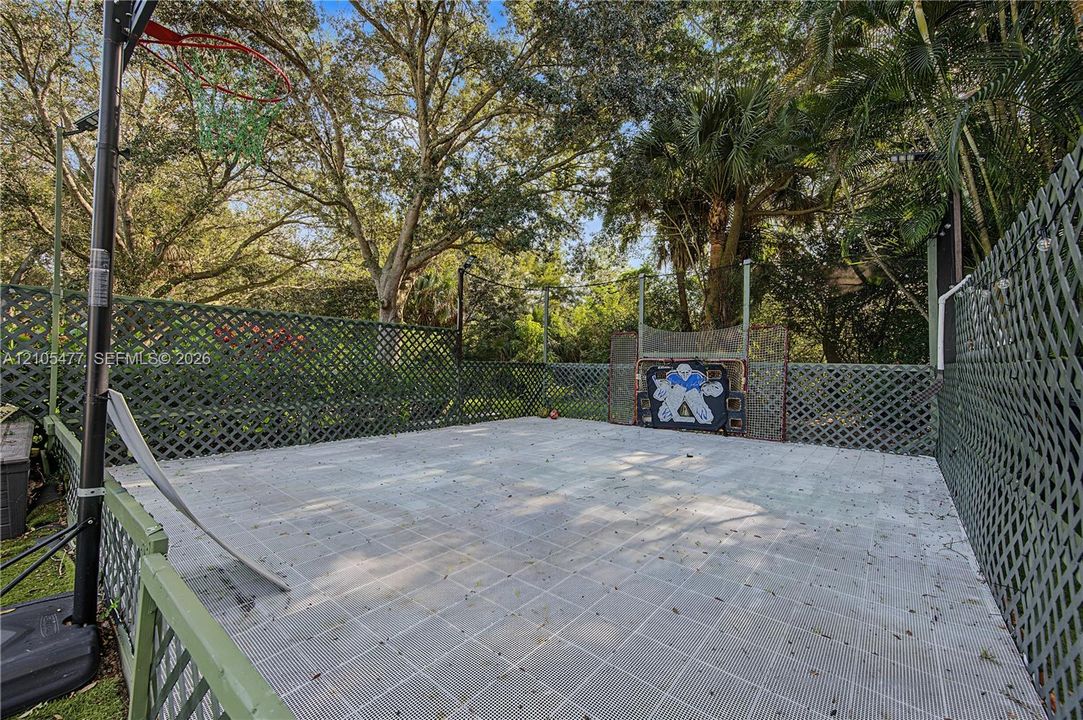 Paved outdoor court featuring a perimeter fence and mature trees