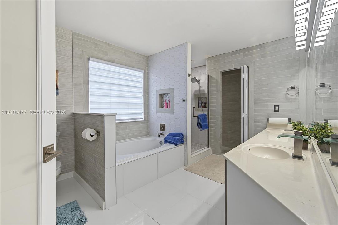 Soaking tub with a window, hexagon tile accent wall, glass-enclosed shower, dual vanity with undermount sinks, and contemporary wall-mounted lighting