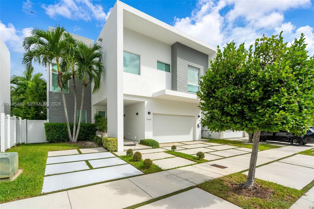 Modern architectural design with a white and gray exterior, integrated garage, concrete and grass paver driveway, and established landscaping