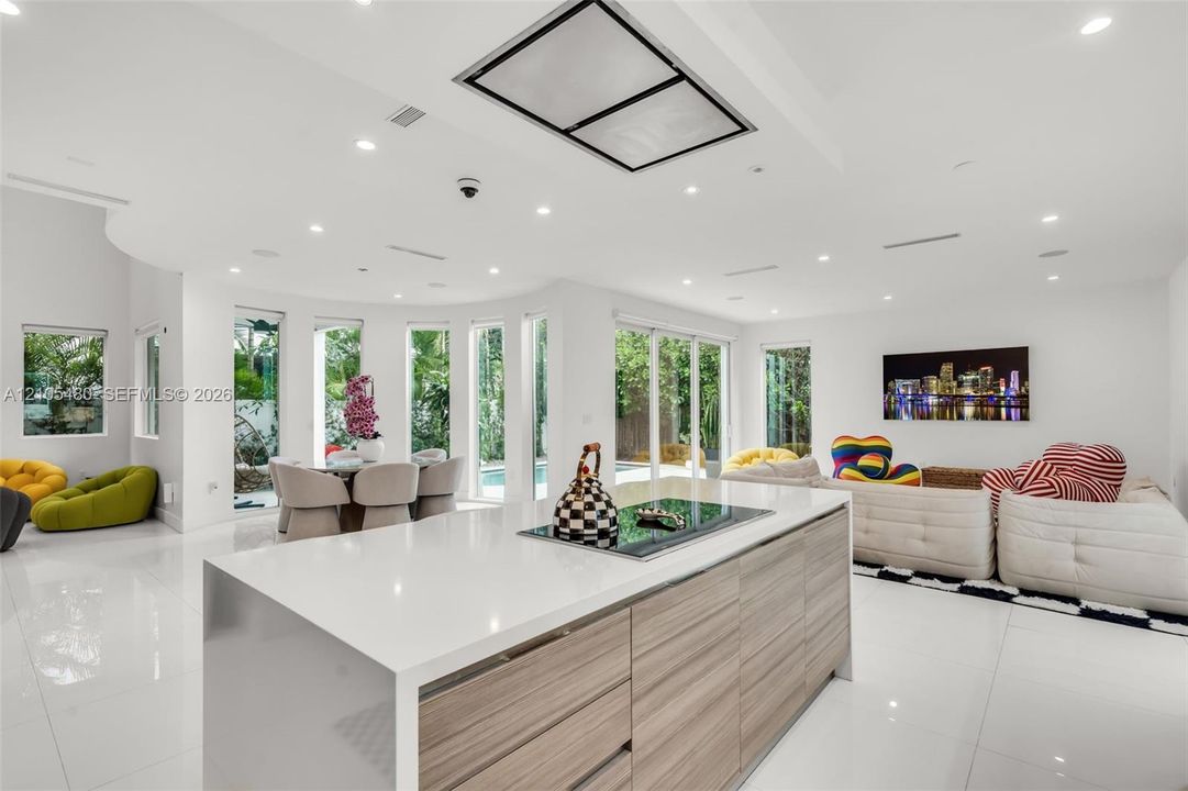 Open-concept living area with white tile flooring, recessed lighting, and a modern island featuring a built-in cooktop and wood-finish cabinetry