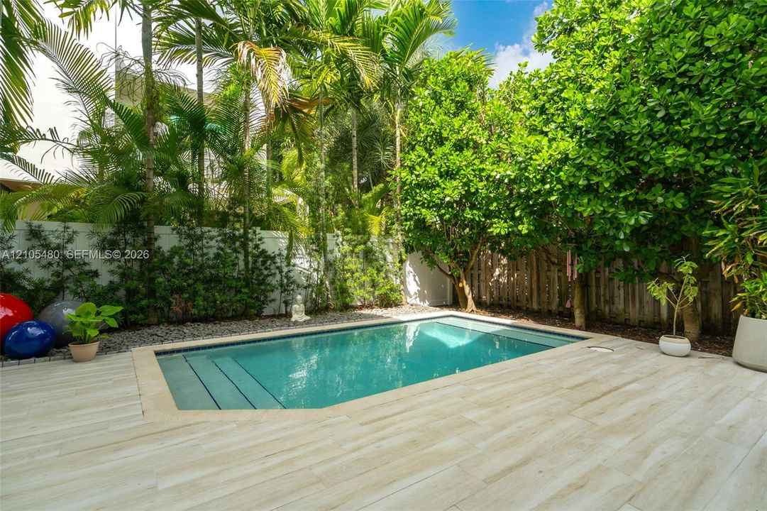 Private backyard oasis featuring a swimming pool with integrated steps, surrounded by light-toned plank decking, lush tropical landscaping, and a privacy fence