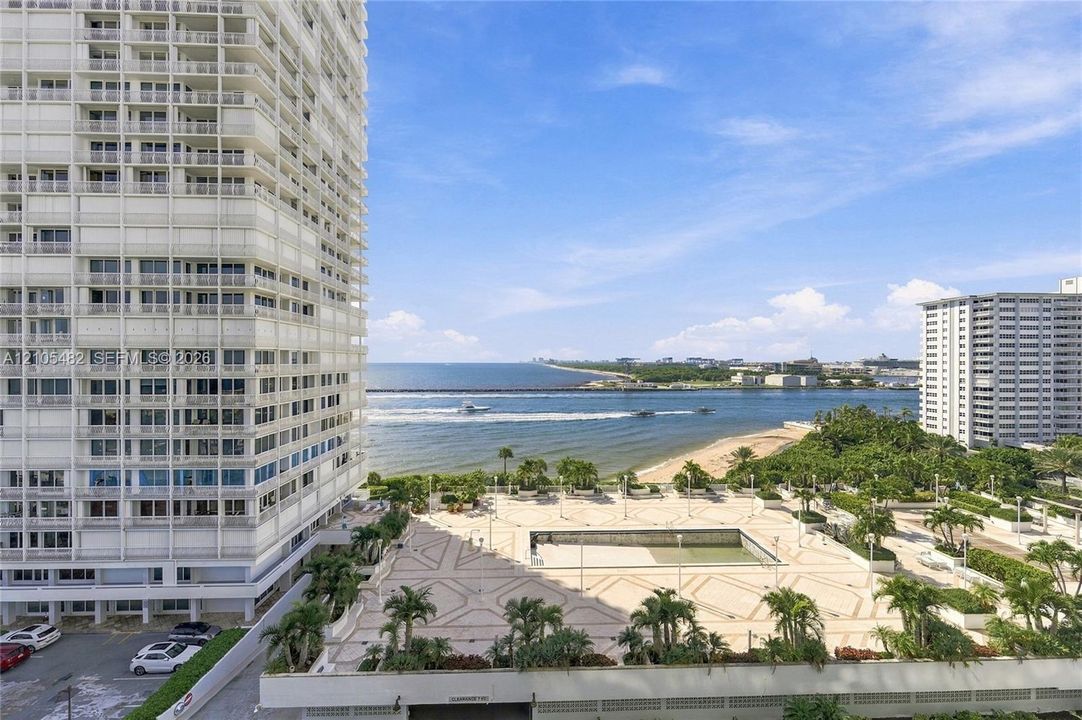 High-rise exterior featuring integrated balconies, stone-tiled pool deck, mature palm landscaping, and expansive ocean and shoreline scenery