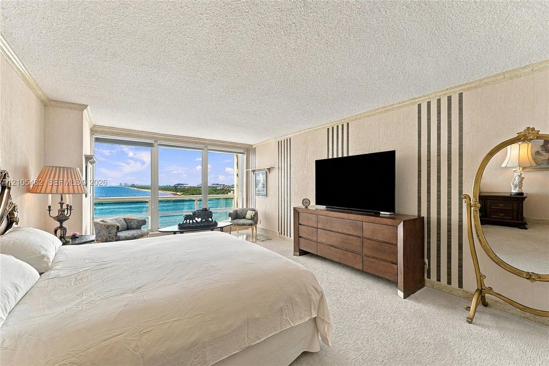Floor-to-ceiling windows with coastal horizon exposure and a private balcony featuring a glass railing, complemented by textured wall treatments and wall-integrated paneling