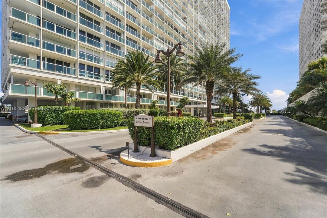 High-rise exterior featuring private balconies with glass railings, landscaped grounds, mature palm trees, and asphalt driveways