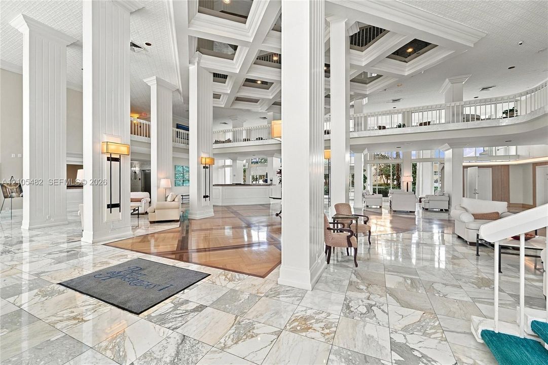 Grand atrium featuring coffered ceilings, polished stone flooring, structural columns with fluted detailing, and a mezzanine level with ornate railings