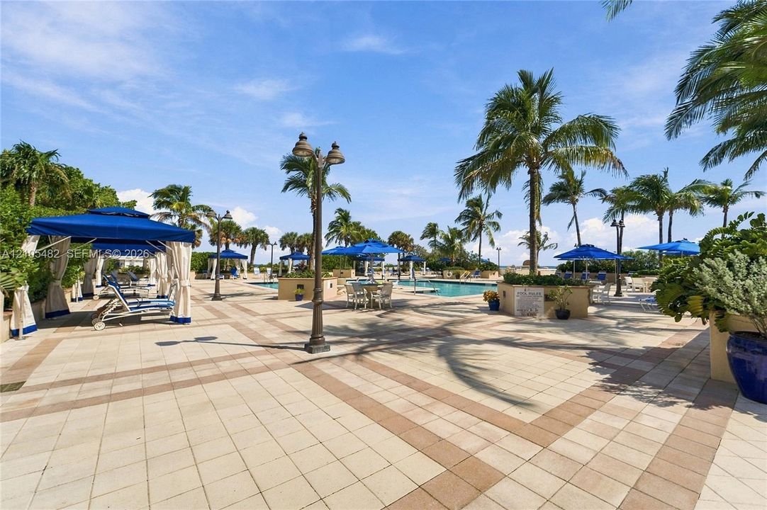 Expansive pool deck featuring light-toned concrete pavers with brick-style accents, an in-ground swimming pool, decorative metal lighting posts, and mature palm trees