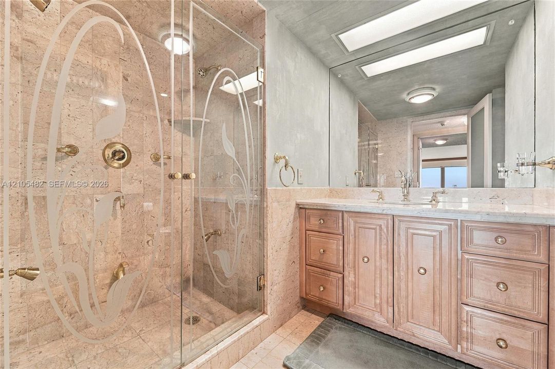Bathroom featuring a walk-in shower with etched glass doors, stone wall cladding, gold-tone fixtures, wood-finish cabinetry, and an expansive wall-to-wall mirror
