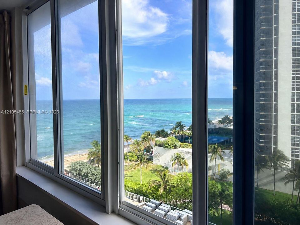Expansive ocean and beach scenery, framed by multiple large windows with white trim, overlooking a tropical landscape