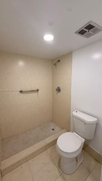 Walk-in shower with tile surround, built-in grab bar, and wall-mounted shower head, accompanied by a porcelain toilet and tile flooring