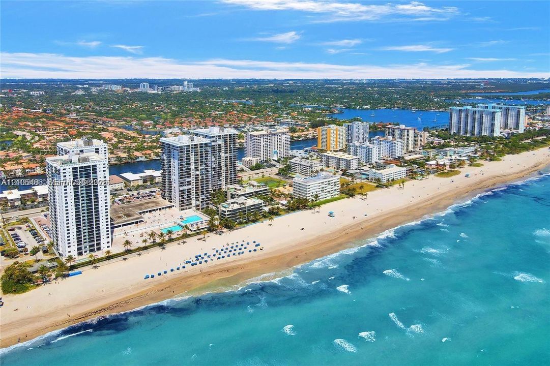 Coastal setting with expansive beach frontage, vibrant blue ocean, and a cityscape backdrop featuring high-rise buildings and a substantial body of water