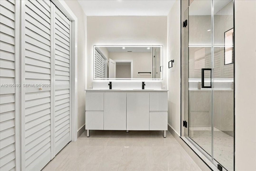 Double vanity with white cabinetry, rectangular vessel sinks, matte black faucets, and a backlit mirror, featuring a walk-in glass-enclosed shower and white louvered closet doors