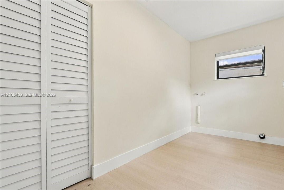 Neutral-toned interior room featuring wood-finish flooring, white louvered closet doors, a horizontal window with integrated roller shade, and wall-mounted utility connections