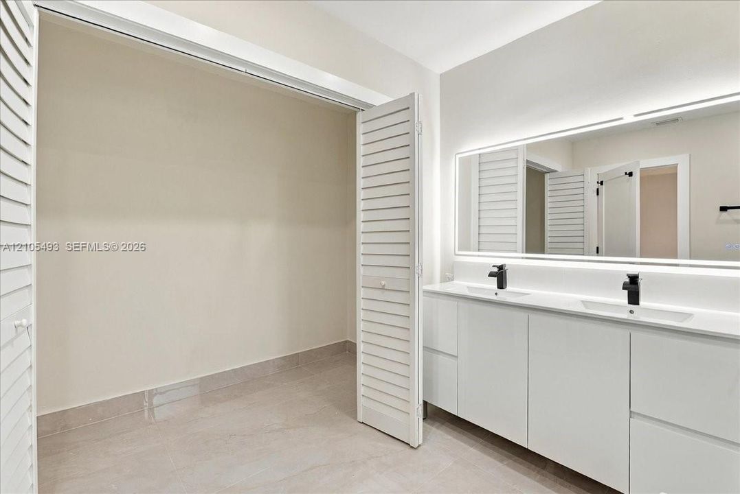 Double vanity with integrated sinks, white cabinetry, matte black hardware, and large illuminated vanity mirror, featuring louvered closet doors and tile flooring