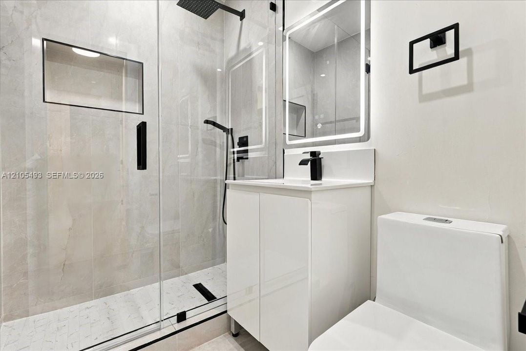 Walk-in shower with stone-finish tiling, rainfall showerhead, and glass enclosure, complemented by a white vanity, integrated LED mirror, and matte black fixtures