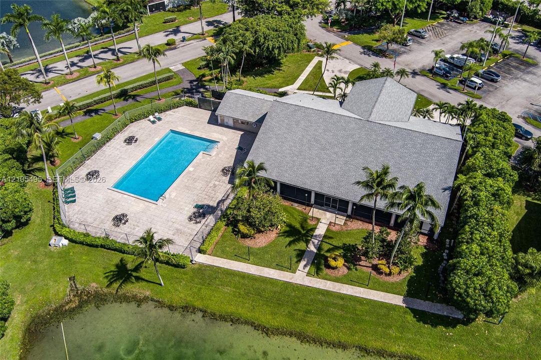 Aerial perspective featuring a rectangular swimming pool, expansive paved patio, landscaped grounds, mature palm trees, and a pond