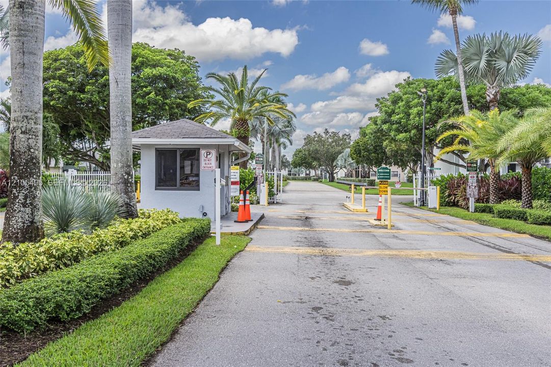 Gated entry featuring a controlled-access guardhouse, paved roadway, ornamental landscaping, and mature palm trees