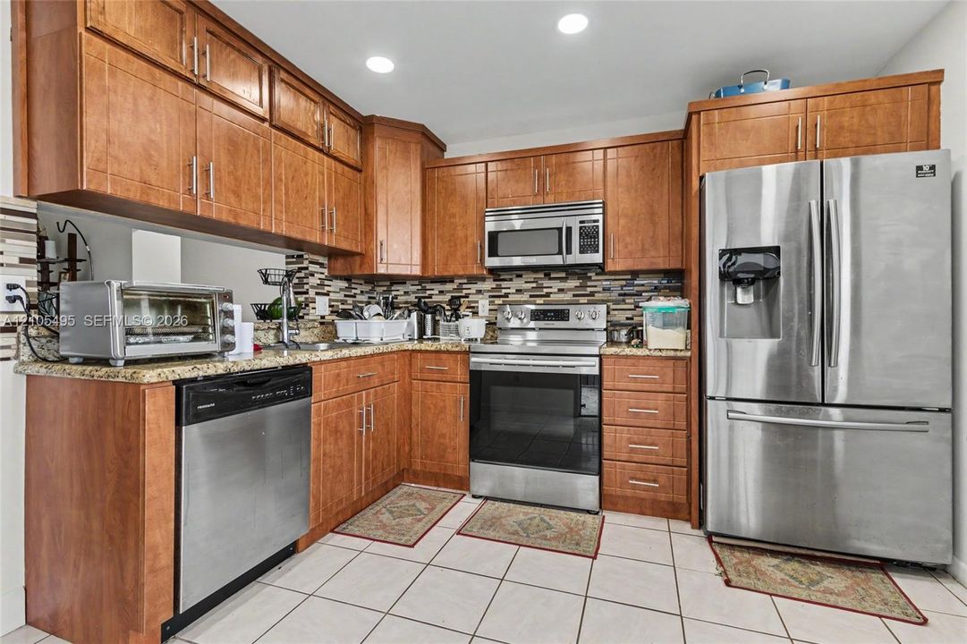 Kitchen featuring shaker-style wood cabinetry, stone countertops, stainless steel appliances, multi-toned tile backsplash, and ceramic tile flooring