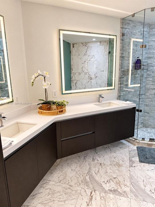 Floating dual vanity featuring dark cabinetry, solid-surface countertops, integrated rectangular sinks, and large, illuminated mirrors, complete with a glass-enclosed shower featuring stone-tiled walls and patterned stone tile flooring