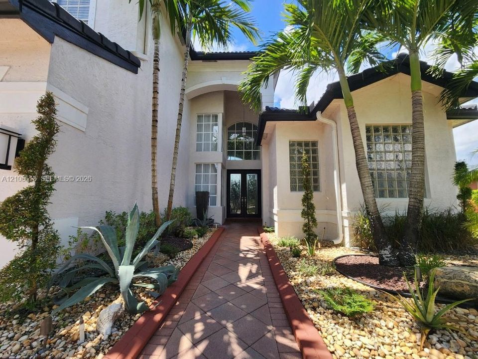 Stucco exterior with a tiled roof, arched entry, double-height windows, and a paved walkway, complemented by mature palm trees and landscaped rock gardens