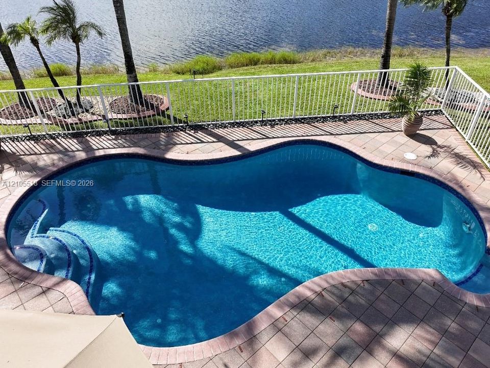 Freeform swimming pool with integrated steps, surrounded by paver decking, and secured by a white perimeter fence