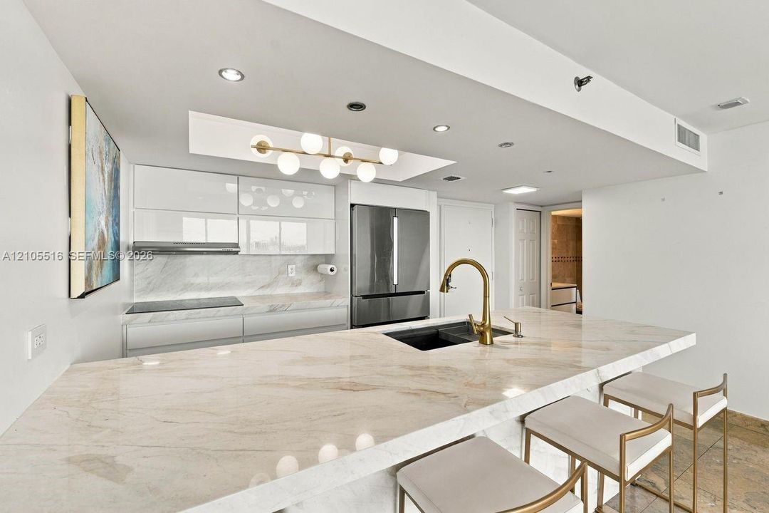 Extended kitchen island with stone countertops, modern pendant lighting, integrated sink with a gold-tone faucet, and white glossy cabinetry